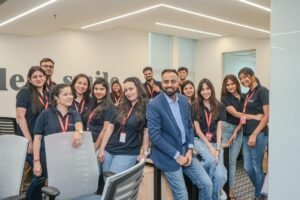 Smiling team posing in an office, several employees in navy shirts with red lanyards surrounding a man in a blue blazer at a desk area.