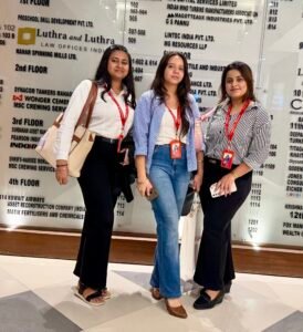 Three women posing together in front of a glass wall with directory listings, wearing red lanyards and carrying bags.