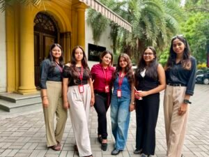 Group of six young women posing outside a yellow building with palm trees, wearing business-casual outfits and red lanyards with ID badges, smiling.