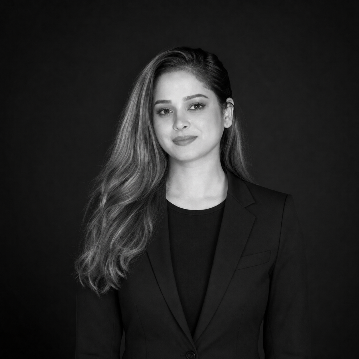 Professional head-and-shoulders portrait of a woman with long wavy hair in a black blazer, against a dark backdrop.