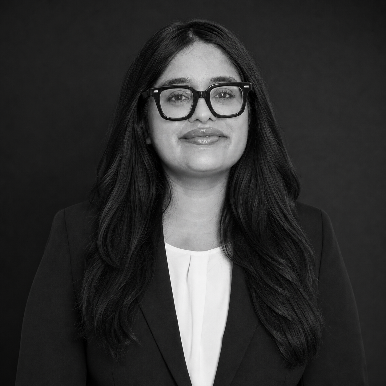 Professional headshot of a woman with long dark hair, black-frame glasses, wearing a blazer and white blouse against a dark background.
