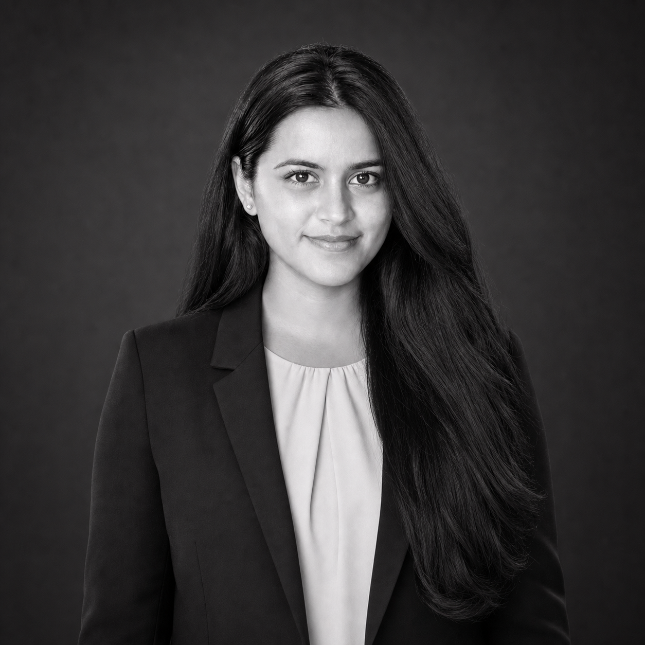 Professional headshot of a young woman with long dark hair in a black blazer, facing the camera, neutral studio background.