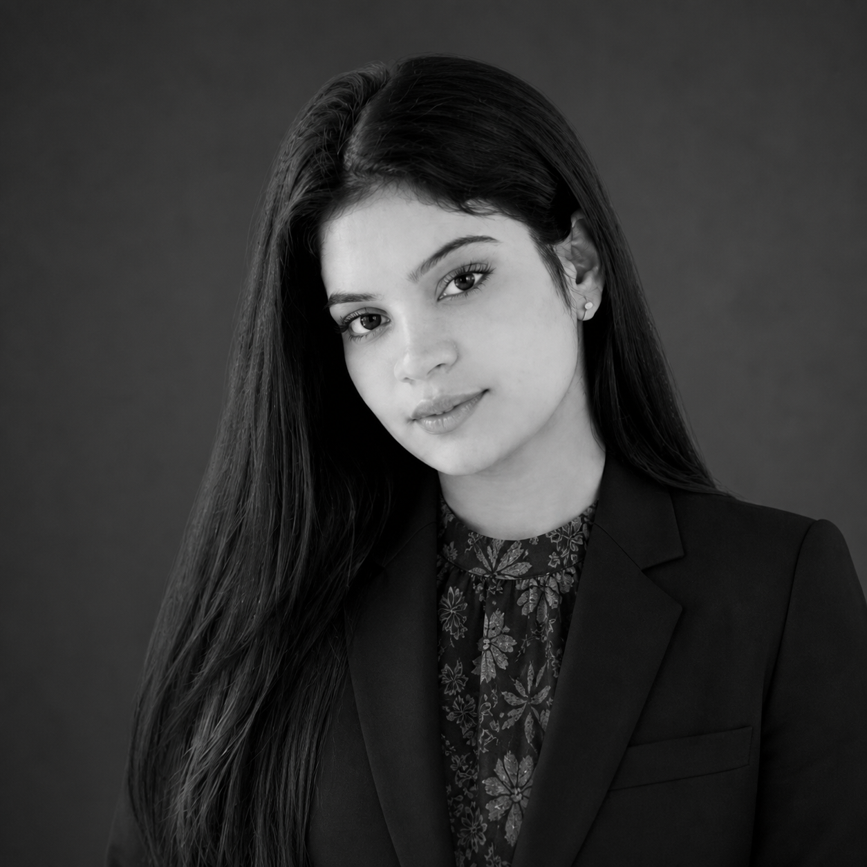 Portrait of a young woman with long dark hair, wearing a black blazer over a floral blouse, in a studio setting.