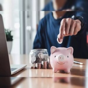 Person places a coin into a pink piggy bank on a wooden desk, with a jar of coins and a laptop nearby.