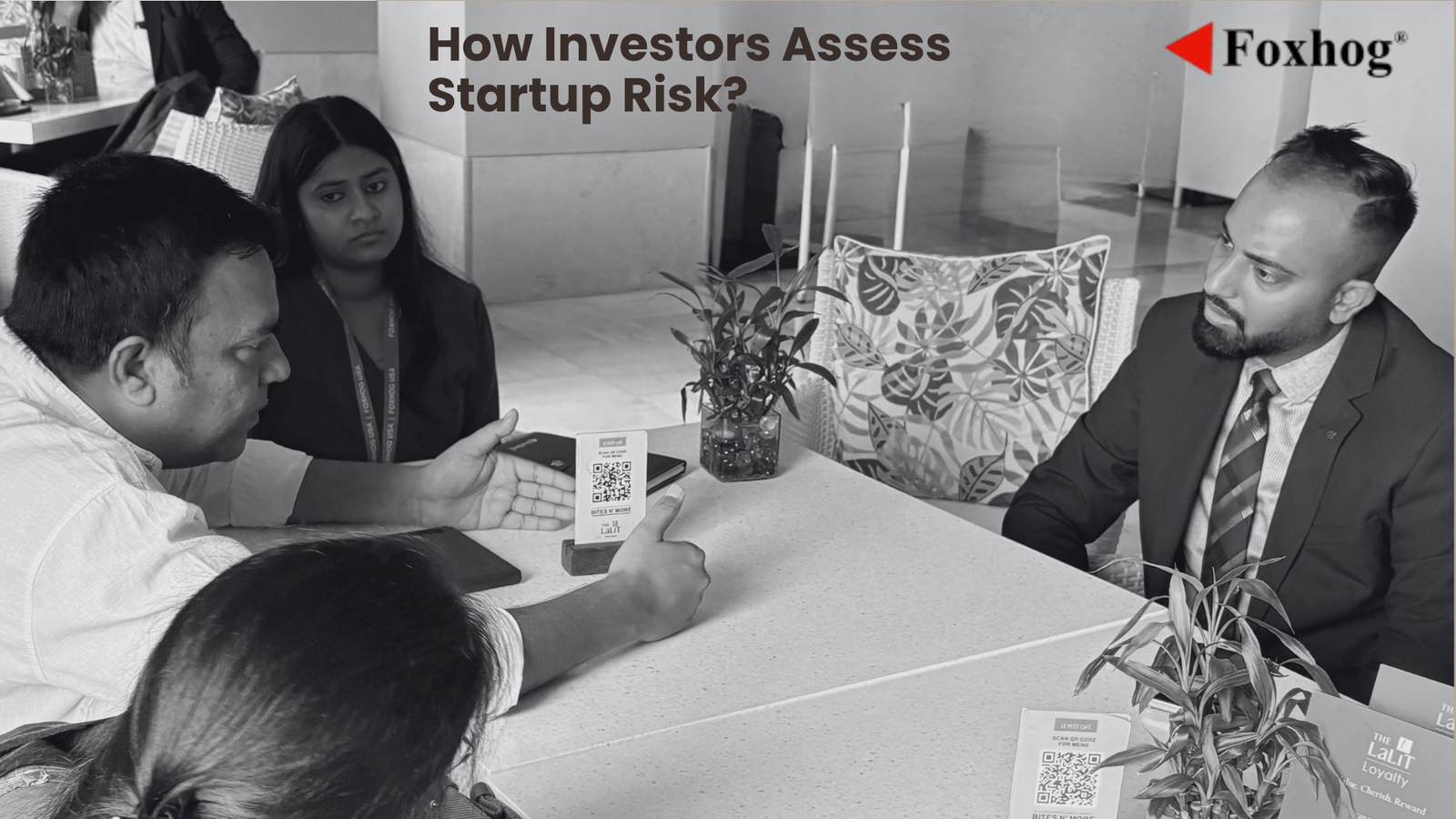 People in a business discussion around a table; a QR code card sits on the table, Foxhog logo top-right, captioned about assessing startup risk.
