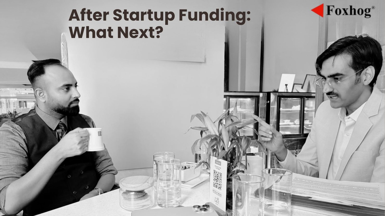 Two men in a cafe discuss post-funding plans over coffee, with a banner reading 'After Startup Funding: What Next?' and Foxhog branding in the corner.